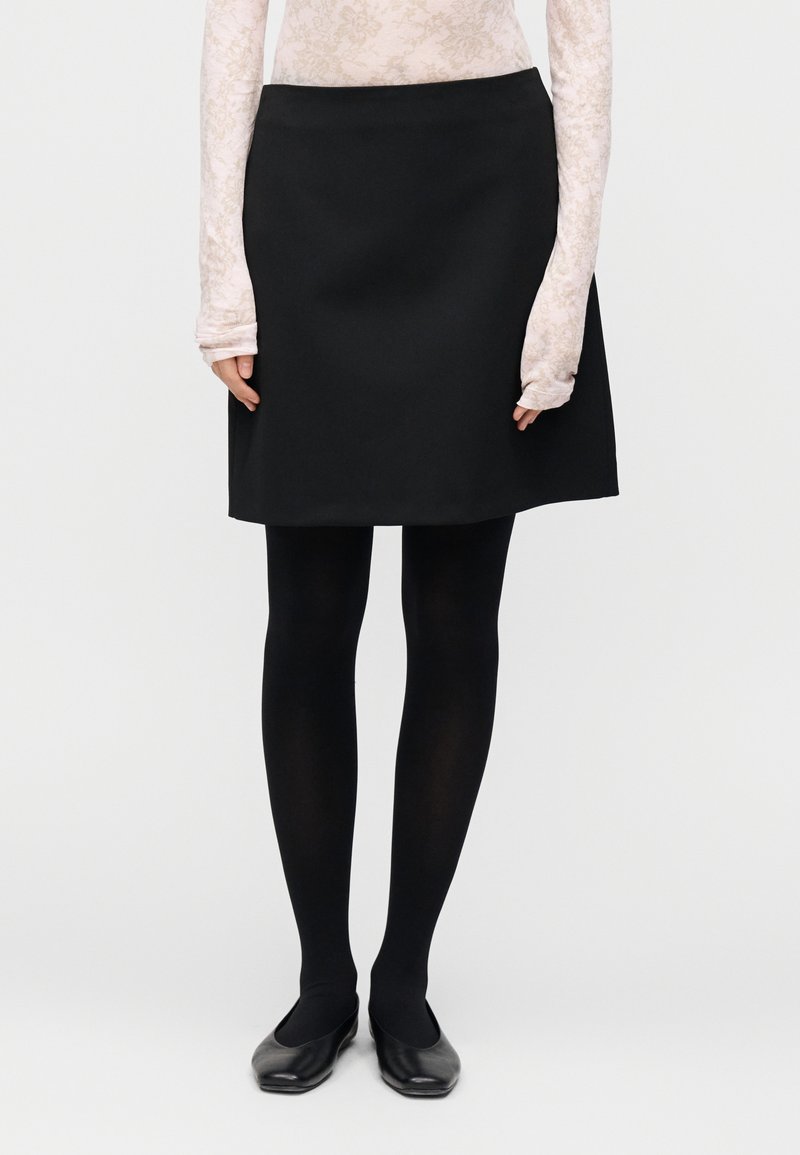 Jupe trapèze noire en tissu lisse, associée à des collants opaques noirs et des chaussures plates noires. Haut à manches longues à motif floral visible en dessous.