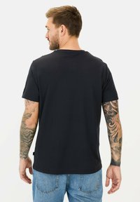 Sort kortærmet t-shirt i et glat materiale, med rund halsudskæring og synlige tatoveringer på armene. Blå jeans er delvist synlige.