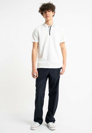 Młody mężczyzna z kręconymi włosami, ubrany w białą koszulkę polo z krótkim rękawem, czarne spodnie i białe trampki, stojący na białym tle.