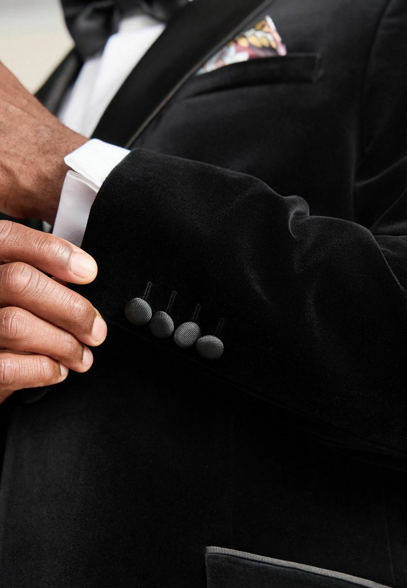 Zwarte fluwelen blazer met een gladde textuur, voorzien van vier gestructureerde zwarte knopen op de manchet en een gestreepte pochet die zichtbaar is in de borstzak.