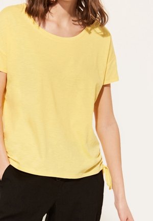 Vrouw die een geel topje met korte mouwen en een zijstrikdetail draagt en zwarte broek, staand tegen een effen lichte achtergrond.
