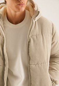 Doudoune en velours côtelé beige avec capuche et doublure sherpa, dotée d'une fermeture éclair et d'une texture côtelée. Portée sur une chemise couleur crème.