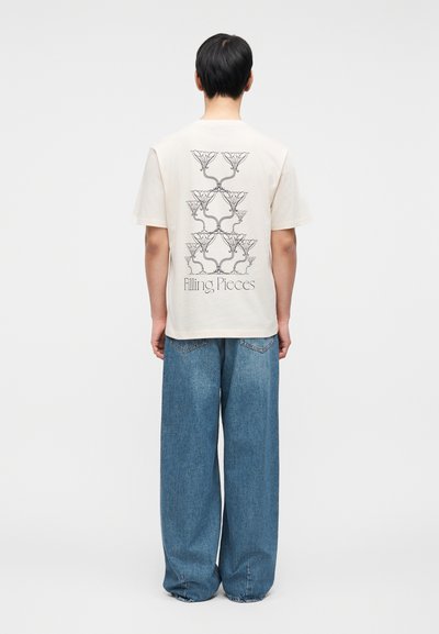 Μπεζ βαμβακερό μπλουζάκι με γραφικό σχέδιο στην πλάτη. Συνδυασμένο με φαρδιά μπλε τζιν παντελόνια, που αναδεικνύουν μια χαλαρή σιλουέτα.