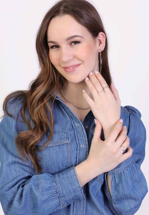 Zilveren sieraden omvatten creolen, gelaagde kettingen en meerdere ringen. Het model draagt een blauwe spijkerblouse met pofmouwen.