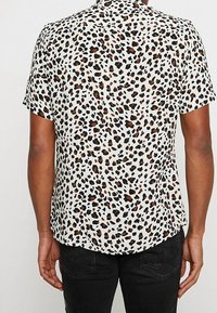 Camicia a maniche corte in tessuto leggero, con uno sfondo bianco decorato da macchie di leopardo nere e marroni, orlo curvo e vestibilità rilassata.