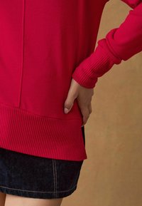 Maglione rosso a maglia con polsini e orlo a coste, abbinato a una gonna di denim scuro. Il materiale sembra morbido e la vestibilità è rilassata.