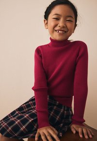 Maglione dolcevita rosso a coste con maniche lunghe, abbinato a una gonna a quadri nei colori blu navy, rosso e bianco; texture liscia, vestibilità casual.