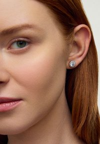 Okrągłe kolczyki wkrętki z przejrzystym, błyszczącym centralnym kamieniem osadzonym w teksturowanym metalowym wykończeniu w srebrnym kolorze. Noszone na uchu modelki.