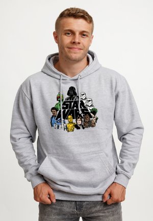 Mand iført grå hættetrøje med farverige Star Wars-figurer og logo, stående mod en ensfarvet hvid baggrund.