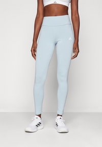 Helle blaue Leggings aus dehnbarem Stoff, mit einem hohen Bund und einem weißen Adidas-Logo auf dem Oberschenkel, kombiniert mit weißen Sportschuhen.