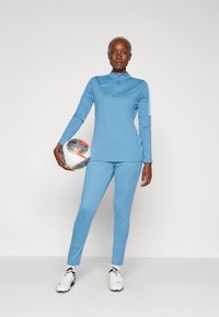 Zila sporta garā piedurkne ar augstu apkakli un atbilstošiem leggingiem, abiem ar Nike logo. Tur rokās rakstainu futbola bumbu.
