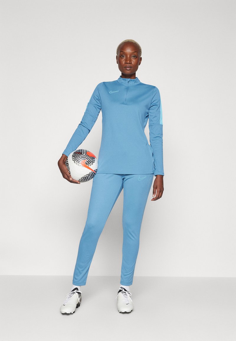 Blaues sportliches Langarmshirt mit hohem Kragen und passenden Leggings, beide mit Nike-Logos. Hält einen gemusterten Fußball.