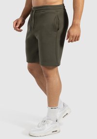 Shorts de algodón verde oliva con cintura elástica, bolsillos laterales y un corte cómodo, combinados con zapatillas deportivas blancas y calcetines hasta el tobillo.