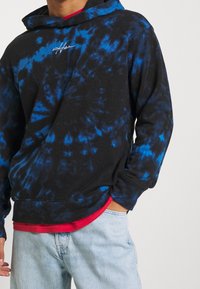 Černá mikina s modrým tie-dye vzorem, s klokaní kapsou, žebrovanými manžetami a malým bílým logem na hrudi.