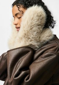 Brązowa skórzana kurtka z dużym, miękkim beżowym futrzanym kołnierzem, o luźnym kroju i zaokrąglonych ramionach.