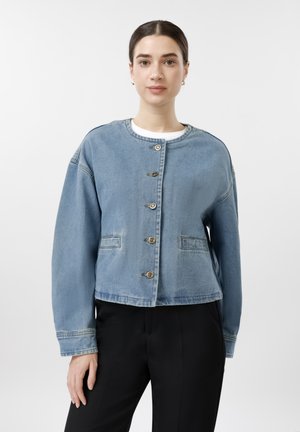 Ung kvinne med mørkt hår iført en blå denimjakke med knapper og svarte bukser, stående foran en ensfarget lys bakgrunn.