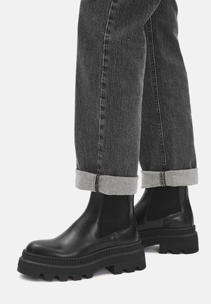 Botines de cuero negro con paneles laterales elásticos, suela gruesa y texturizada, y acabado mate, combinados con vaqueros grises remangados.