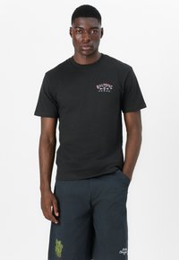 Svart bomulls t-skjorte med kort erme, med en liten fargerik logo på venstre brystområde. Modellen har på seg grå shorts med grønne detaljer.