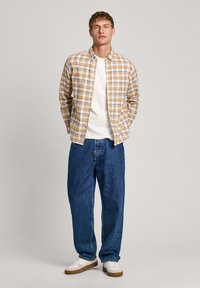 Camicia plaid abbottonata in crema, giallo e marrone, sopra una maglietta bianca. Indossata con jeans in denim blu larghi e sneakers bianche con suole marroni.