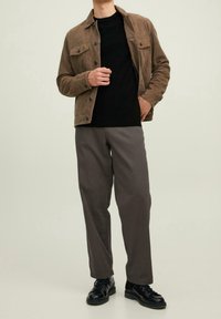 Veste en daim marron avec poches avant, t-shirt noir, pantalon gris ample et chaussures noires à lacets, sur un fond clair.