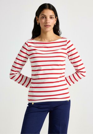 Vrouw met lang donker haar die gouden oorbellen draagt, een wit shirt met lange mouwen met rode horizontale strepen en een marineblauwe broek met gouden knopen.