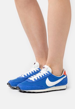 Niebieskie sneakersy z siateczki i zamszu z białymi sznurówkami oraz logo Nike. Teksturowana cholewka, czerwony akcent na pięcie i czarna gumowa podeszwa.