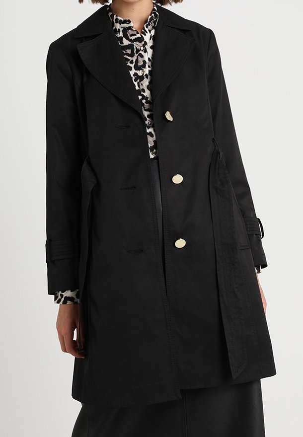 Femme portant un trench-coat noir avec trois boutons dorés sur un chemisier imprimé léopard, debout devant un fond clair uni.