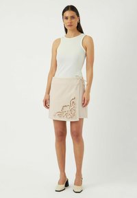 Jupe portefeuille beige avec un motif de broderie en dentelle florale sur le côté gauche, associée à un débardeur blanc ajusté et des chaussures à talons carrés.