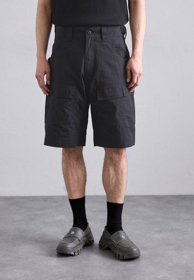 Homme portant un short cargo noir, des chaussettes noires et des chaussures à enfiler épaisses et sombres, se tenant sur un sol gris contre un mur uni.