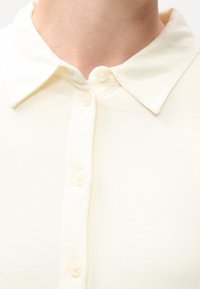 Gros plan d'une personne portant une chemise crème boutonnée avec un col classique, montrant le cou et la partie supérieure de la poitrine.