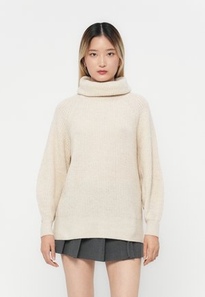 Beige strikkegenser med høy, brettet krage og lange, litt puffete ermer. Strukturfabric og avslappet passform over et grått skjørt.