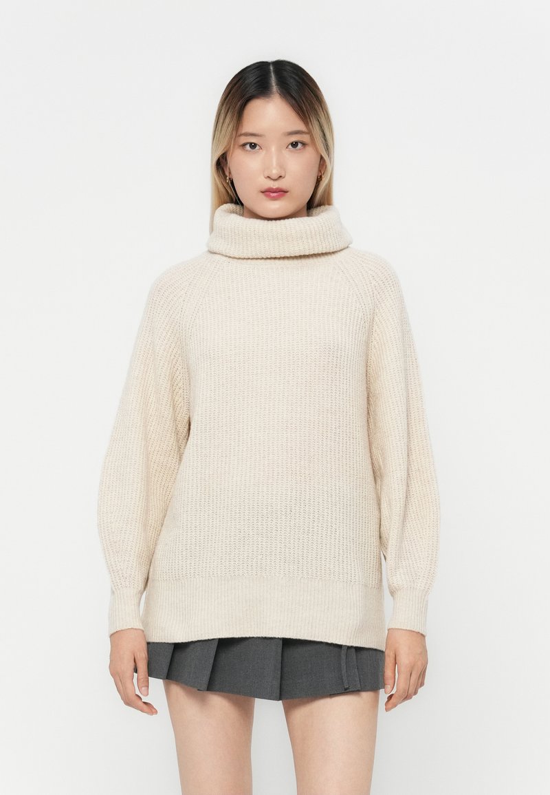 Beiger gestrickter Pullover mit hohem, umgeschlagenem Kragen und langen, leicht gepufften Ärmeln. Strukturierter Stoff und lockere Passform über einem grauen Rock.