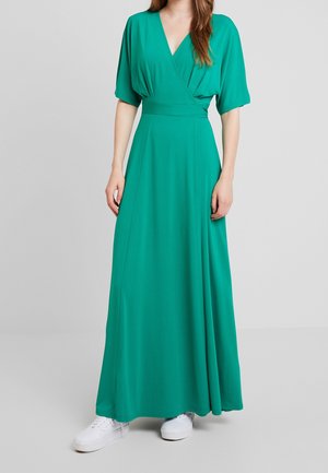 Kobieta w długiej, sięgającej podłogi sukience w kolorze teal, z krótkimi rękawami i dekoltem w serek, w białych trampkach, stojąca na jednolitym tle.