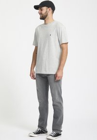 Gray short-sleeve t-shirt featuring a small logo, paired with faded gray jeans and black sneakers with white soles. The model wears a black cap.