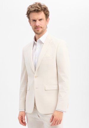 Man met lichtbruin haar en baard, gekleed in een crèmekleurig pak en wit overhemd, staand tegen een effen witte achtergrond.