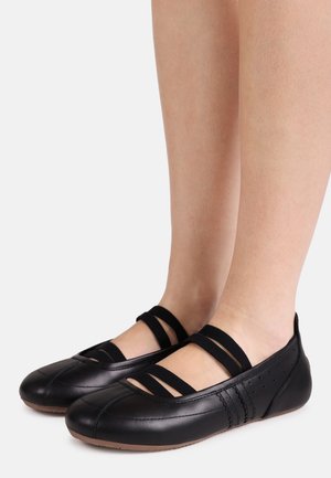 Füße, die schwarze Ballettschuhe aus Leder mit drei elastischen Riemen und braunen Sohlen auf hellem grauem Hintergrund tragen.