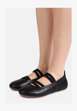 Voeten met zwarte lederen ballerina's met drie elastische bandjes en bruine zolen op een lichtgrijze achtergrond.