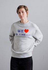 Sivá mikina s okrúhlym výstrihom, so výrazným námorníckym modrým a červeným nápisom "MILUJEME NEW YORK CITY." Voľný strih s rebrovanými manžetami a lemom.