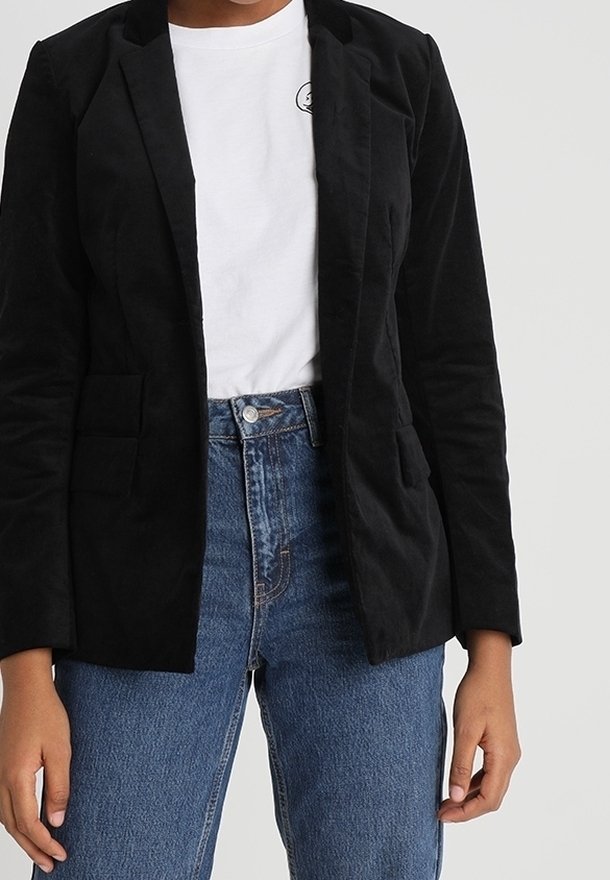 Personne portant un blazer noir par-dessus un t-shirt blanc et un jean bleu taille haute, debout devant un arrière-plan clair uni.