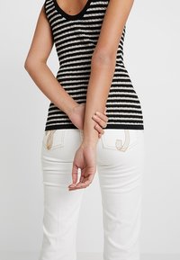 Schwarz-weiß gestreifes gestricktes Tanktop mit rundem Halsausschnitt, kombiniert mit weißen Jeans, die dekorative Nähten an den Gesäßtaschen aufweisen.