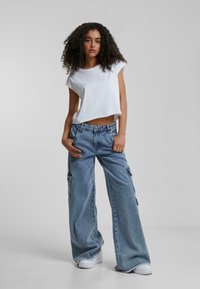 Fehér, rövid ujjú, cropped póló, amelyet világoskék, bő szárú farmerral viselnek, oldalsó zsebekkel és szakadt szegéllyel, fehér sportcipővel kiegészítve.