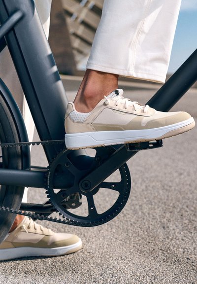 Béžové sportovní boty s síťovými akcenty a světle šedou podrážkou, umístěné na pedálu jízdního kola na texturovaném povrchu.