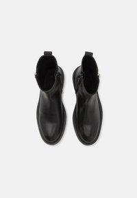 Botine negre din piele, cu o textură netedă, vârf rotund, închidere cu fermoar și maner pentru a fi ușor de încălțat. Talpă plată cu detalii de cusătură subtile.