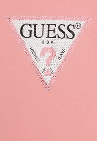 T-shirt rose avec un logo blanc triangulaire, du texte noir et des strass. Le design comprend le nom de la marque et un point d'interrogation.