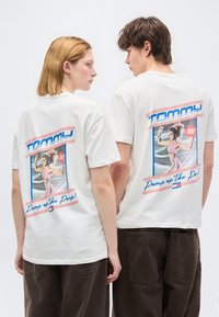 Δύο άτομα φορούν λευκά μπλουζάκια με γραφικά, με εικόνα ρετρό γυμναστικής και το κείμενο «Tommy Pump up The Pwr!» στην πλάτη.