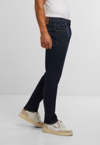 Tumši zils denima bikses ar taisnleņķa piegriezumu, ar brūnu ādas uzšuvi aizmugurē, smalku šuvēm un sānu kabatām.