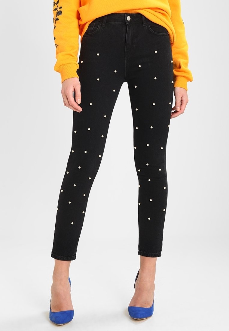 Femme portant un jean skinny noir avec des clous blancs en forme de perles, un sweat-shirt orange vif et des chaussures à talons bleues, debout sur un fond blanc.