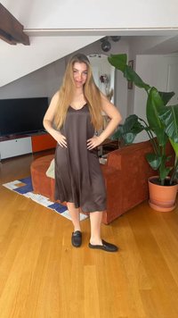 Γυναίκα με μακριά ξανθά μαλλιά που φοράει σκούρο νυχτικό και μαύρα παπούτσια χωρίς κορδόνια, στέκεται σε ένα σαλονάκι με ξύλινο δάπεδο και μεγάλο πράσινο φυτό.