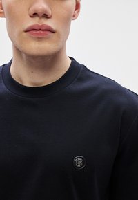 Μπλε ναυτικό βαμβακερό φούτερ με στρογγυλή λαιμόκοψη, που διαθέτει μικρό, κυκλικό λογότυπο στο αριστερό μέρος του στήθους. Ομαλή υφή και εφαρμοστή σχεδίαση.