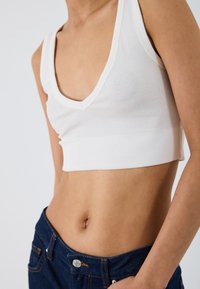 Fehér bordás, cropped tank top V-nyakkal, testhez simuló kialakítással. Sötét farmernadrággal párosítva, amely egy laza, réteges megjelenést mutat.